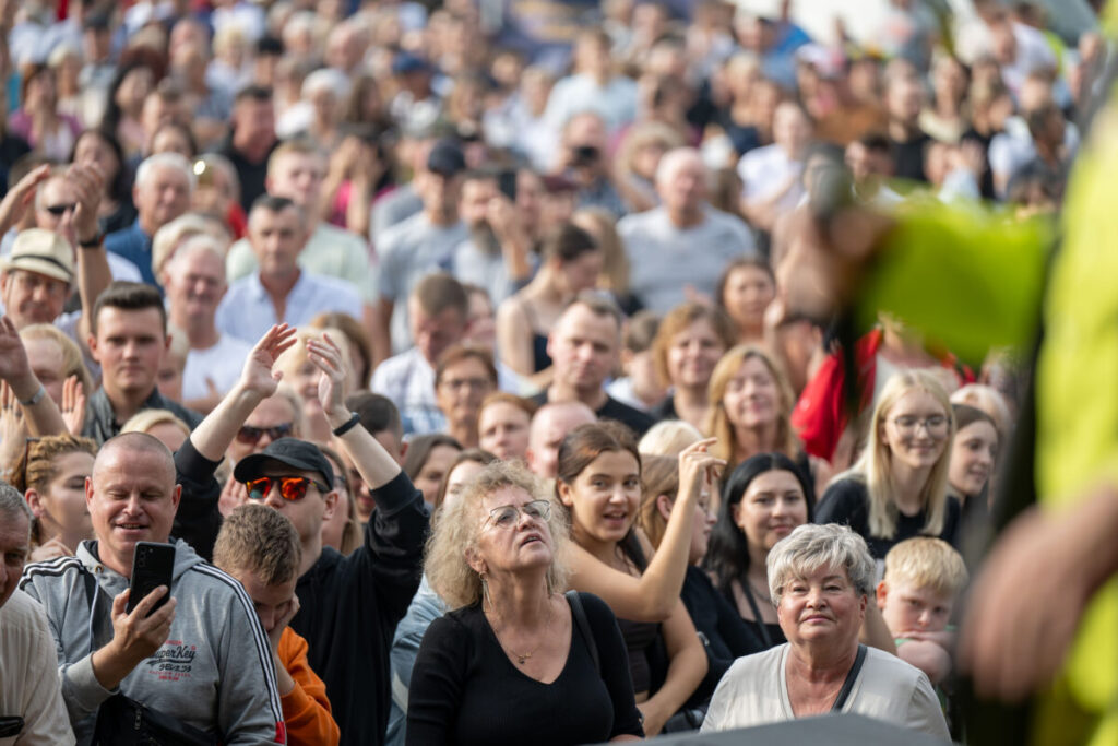 Zdjęcie przedstawia tłum ludzi na imprezie plenerowej. Kolory są jasne, przeważają odcienie bieli i szarości. W tle widać zróżnicowane twarze pełne entuzjazmu.