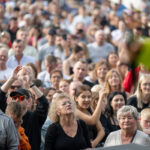 Zdjęcie przedstawia tłum ludzi na imprezie plenerowej. Kolory są jasne, przeważają odcienie bieli i szarości. W tle widać zróżnicowane twarze pełne entuzjazmu.