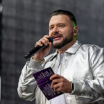 Osoba w srebrnej kurtce trzyma mikrofon i notatki na scenie. Tło jest ciemne, rozmyte. Ma krótko przycięte włosy i brodę.