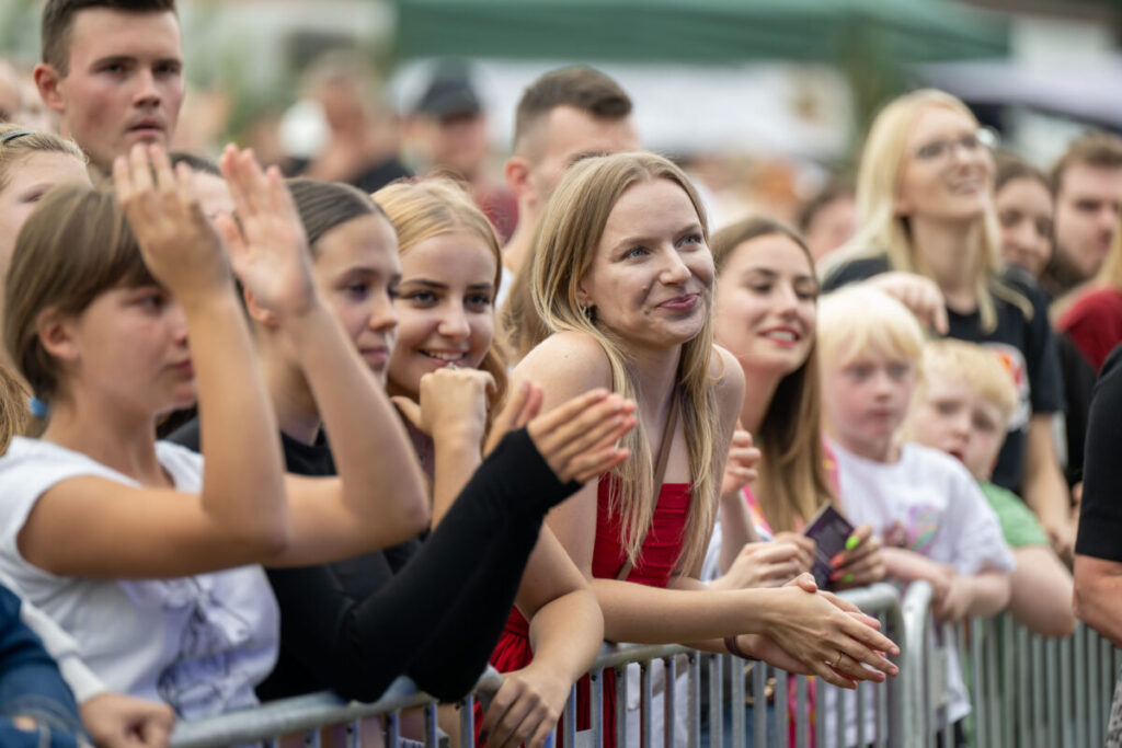 Grupa osób stoi za barierką na koncercie na świeżym powietrzu. Widać uśmiechnięte twarze, radosną atmosferę i różnorodne kolory ubrań. Tło jest rozmyte, sugerując środowisko festiwalowe.