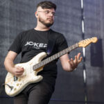 Mężczyzna w czarnej koszulce gra na elektrycznej gitarze. Tło jest szare i matowe. Instrument ma kremowy kolor i widoczne ślady zużycia.