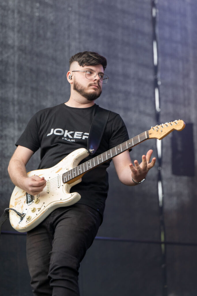 Mężczyzna w czarnej koszulce gra na elektrycznej gitarze. Tło jest szare i matowe. Instrument ma kremowy kolor i widoczne ślady zużycia.