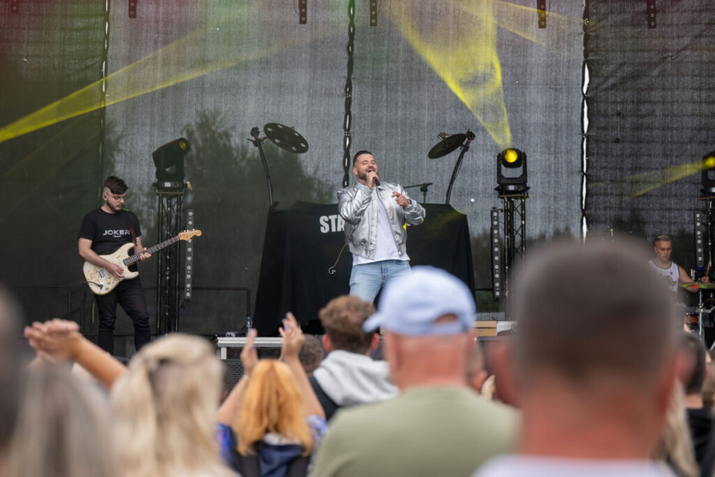 Na scenie mężczyzna śpiewa, ubrany w srebrną kurtkę. Obok gitarzysta, w tle ciemna scenografia i kolorowe światła. Publiczność rozentuzjazmowana, widać uniesione ręce.