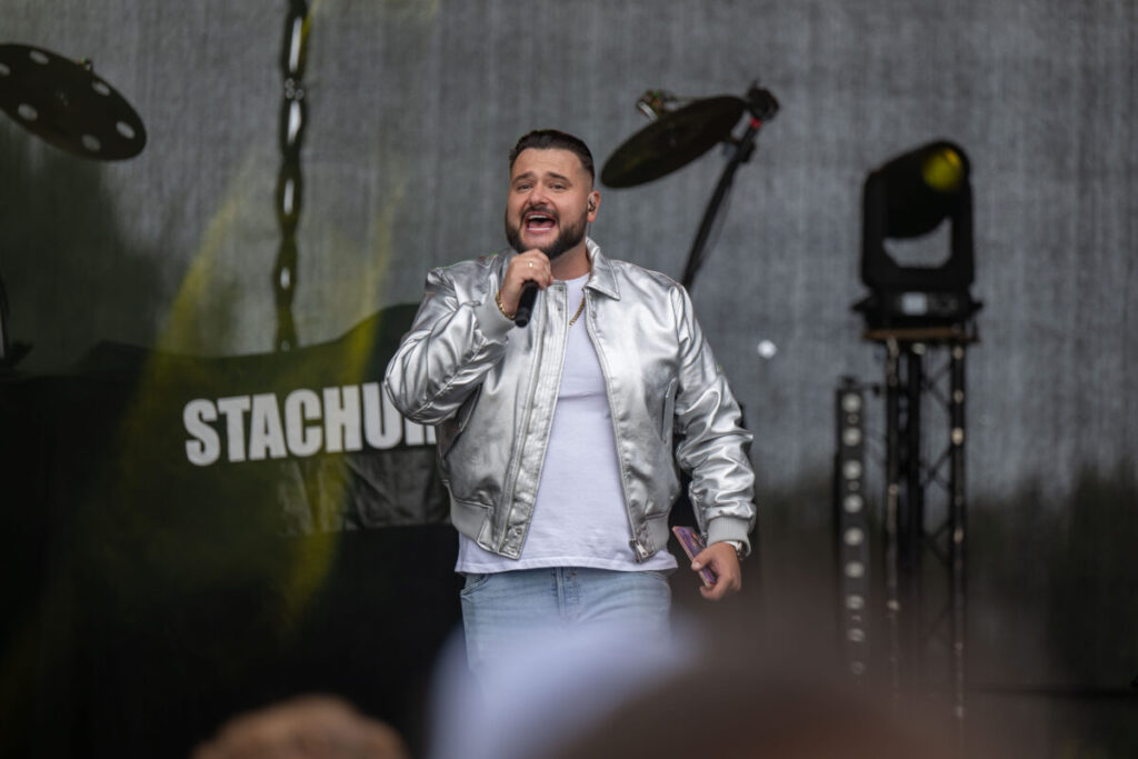 Mężczyzna w srebrnej kurtce śpiewa na scenie. Tło jest ciemne z widocznymi światłami sceny. Na banerze napis STACHU.