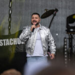 Mężczyzna w srebrnej kurtce śpiewa na scenie. Tło jest ciemne z widocznymi światłami sceny. Na banerze napis STACHU.