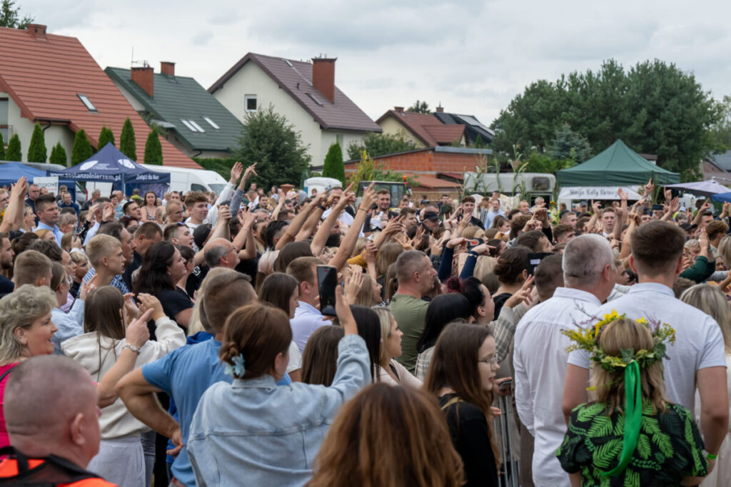 Tłum zbierający się na zewnątrz z podniesionymi rękami. W tle namioty i domy, przeważają kolory zielony i czerwony. Dzień jest pochmurny.