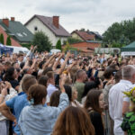 Tłum zbierający się na zewnątrz z podniesionymi rękami. W tle namioty i domy, przeważają kolory zielony i czerwony. Dzień jest pochmurny.