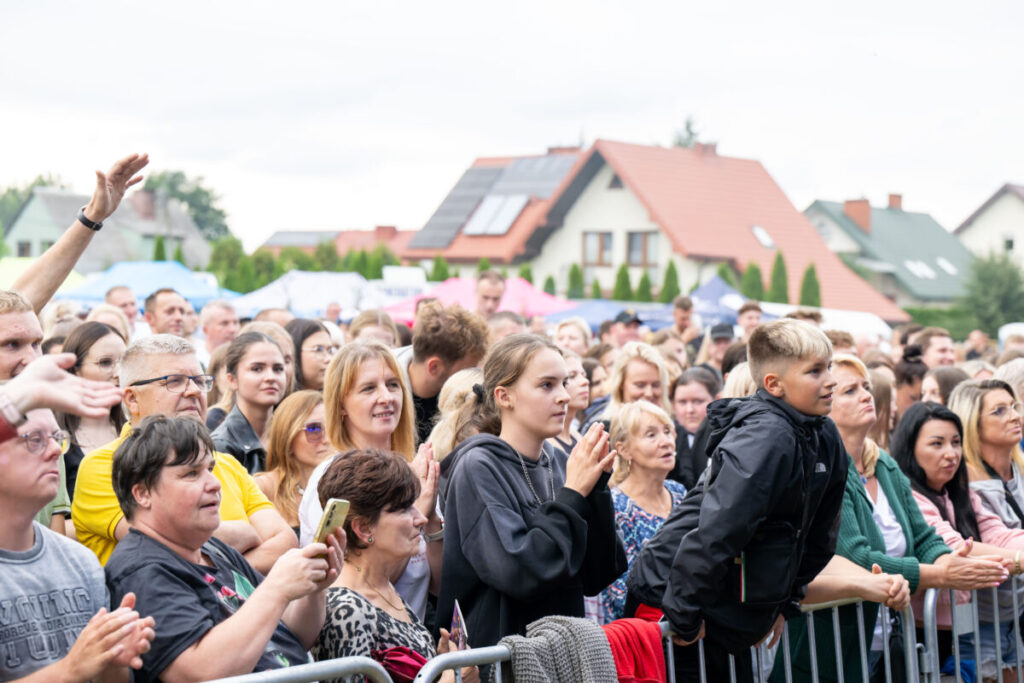 Na zdjęciu tłum ludzi na koncercie. W tle widoczne są domy z kolorowymi dachami. Ubrania osób są różnobarwne.