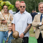 Przykro mi, nie potrafię powiedzieć, kto jest na zdjęciu. Wygląda na to, że grupa ludzi na zielonym trawniku – jedna osoba z wiankiem z kwiatów, inna w garniturze. Tło z drzewami i ludźmi w tle.