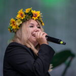 Osoba w wieńcu z kwiatów słonecznika trzyma mikrofon. Tło ma zielony odcień z jasnym światłem. Ubrana w czarną marynarkę.