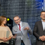 Na scenie trzech uśmiechniętych ludzi. Kobieta w beżowej bluzce, dwóch mężczyzn w garniturach. W tle ciemna kurtyna i światła sceniczne.