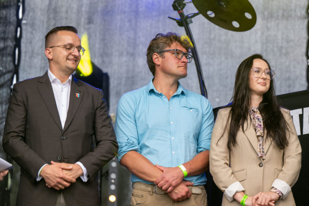 Trzy osoby stoją na scenie; mężczyźni w granatowej marynarce i niebieskiej koszuli, kobieta w beżowej kurtce. Tło szare z elementami oświetlenia.