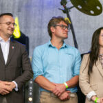 Trzy osoby stoją na scenie; mężczyźni w granatowej marynarce i niebieskiej koszuli, kobieta w beżowej kurtce. Tło szare z elementami oświetlenia.