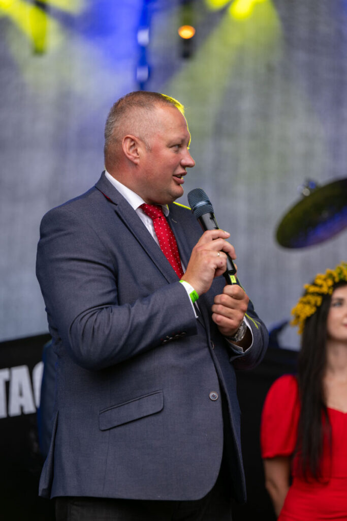 Mężczyzna w garniturze i czerwonym krawacie mówi do mikrofonu. W tle rozmyte światła sceniczne i kobieta w czerwieni z wiankiem. Dominują kolory niebieski, czerwony i żółty.