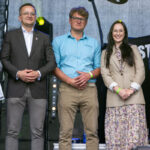 Trzy osoby stoją na scenie. Mężczyzna w garniturze po lewej, mężczyzna w niebieskiej koszuli pośrodku i kobieta w sukience we wzory po prawej. Tło jest szare, z elementem technicznym za nimi.