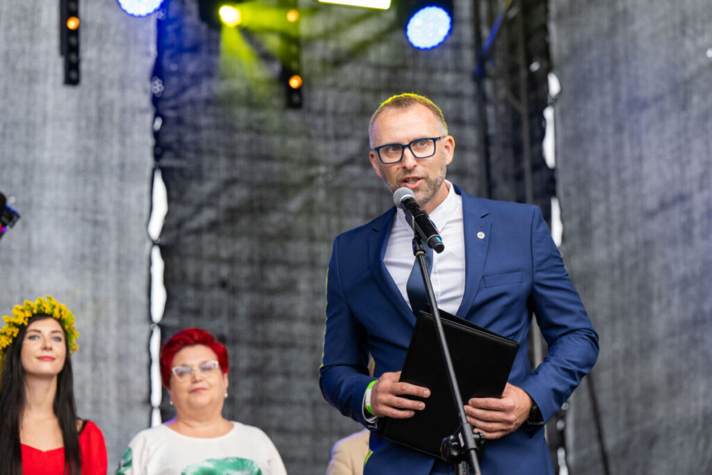 Mężczyzna w niebieskim garniturze przemawia przez mikrofon na scenie. Tło jest ciemne z kolorowymi światłami. Obok stoją dwie kobiety.