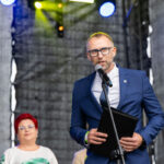 Mężczyzna w niebieskim garniturze przemawia przez mikrofon na scenie. Tło jest ciemne z kolorowymi światłami. Obok stoją dwie kobiety.