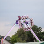 Na zdjęciu widać jasną karuzelę Monster z pasażerami w ruchu. Kolory to głównie fiolet i biały. W tle drzewa i domy.