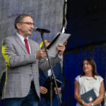 Mężczyzna w szarej marynarce przemawia przy mikrofonie. Obok stoi kobieta w białej sukience z teczką. Tło jest ciemne, scena jest dobrze oświetlona.