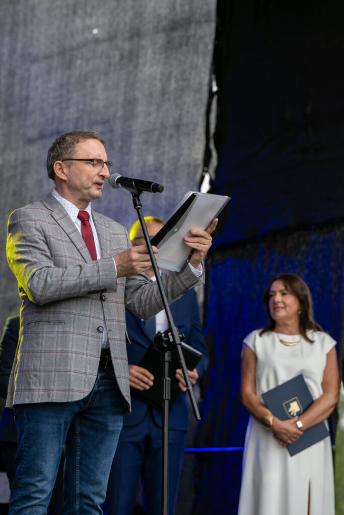 Mężczyzna w szarej marynarce przemawia przy mikrofonie. Obok stoi kobieta w białej sukience z teczką. Tło jest ciemne, scena jest dobrze oświetlona.