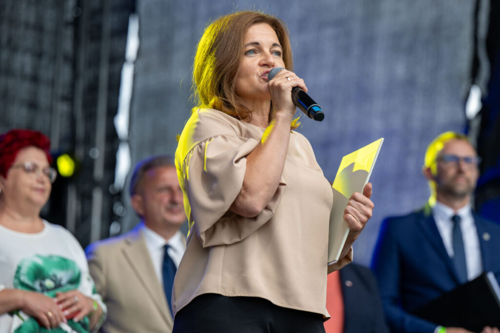 Na zdjęciu kobieta w beżowej bluzce przemawia do mikrofonu, trzymając dokumenty. W tle grupa osób formalnie ubranych. Scena ma szare tło.