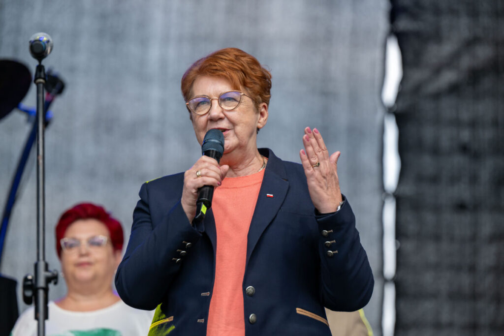 Na zdjęciu kobieta w ciemnej marynarce z mikrofonem, w tle szare tło. Ma krótkie, rude włosy. Obok stoi inna osoba.
