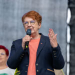 Na zdjęciu kobieta w ciemnej marynarce z mikrofonem, w tle szare tło. Ma krótkie, rude włosy. Obok stoi inna osoba.