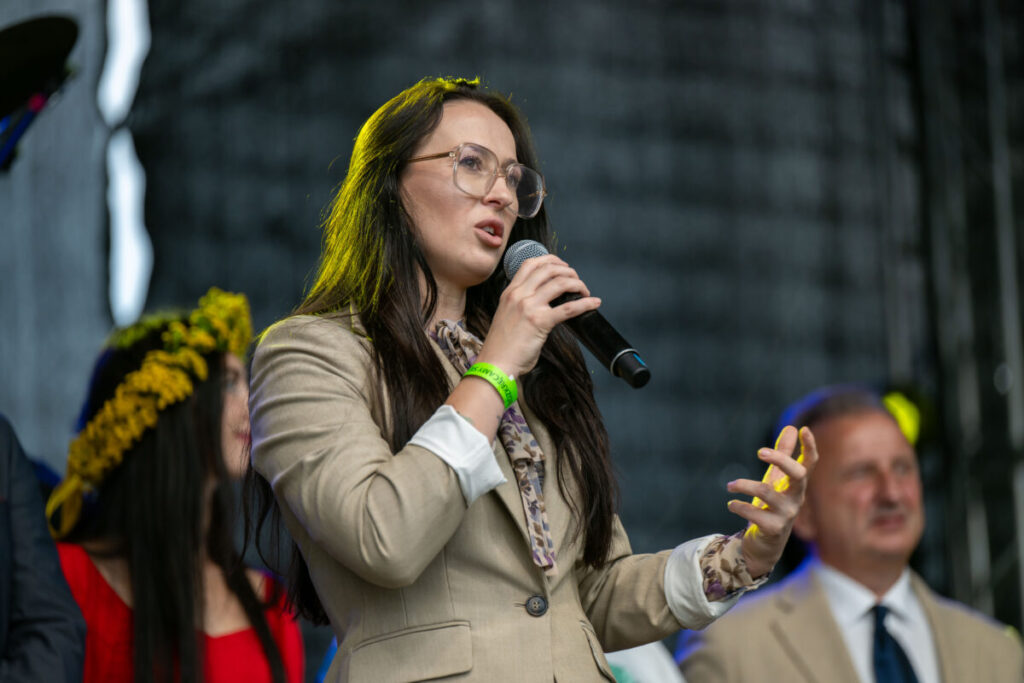 Nie wiem, kim są osoby na zdjęciu. Młoda kobieta w okularach, w beżowym garniturze, mówi do mikrofonu. W tle ciemne tło i inne, nieznane postacie.