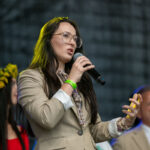 Nie wiem, kim są osoby na zdjęciu. Młoda kobieta w okularach, w beżowym garniturze, mówi do mikrofonu. W tle ciemne tło i inne, nieznane postacie.