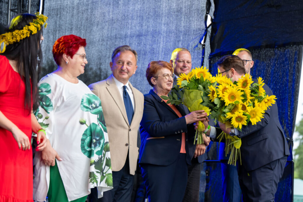 Na scenie grupa ludzi, w środku kobieta z bukietem słoneczników. Kolorowe stroje, niebieskie tło. Tło sceniczne.