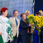 Na scenie grupa ludzi, w środku kobieta z bukietem słoneczników. Kolorowe stroje, niebieskie tło. Tło sceniczne.