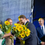 Osoby na zdjęciu wymieniają bukiety słoneczników. Kobieta w białej bluzce z zielonym wzorem. Tło jest ciemnoniebieskie.