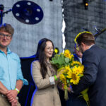 Na scenie kilka osób; jedna z kobiet trzyma bukiet słoneczników. Mężczyzna w garniturze wręcza kwiaty. Tło szare, industrialne.