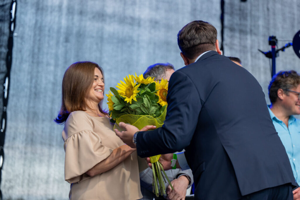 Na zdjęciu kobieta z uśmiechem odbiera bukiet słoneczników od mężczyzny. W tle ciemnoszara kurtyna. Obok stoją osoby w niebieskich koszulach.