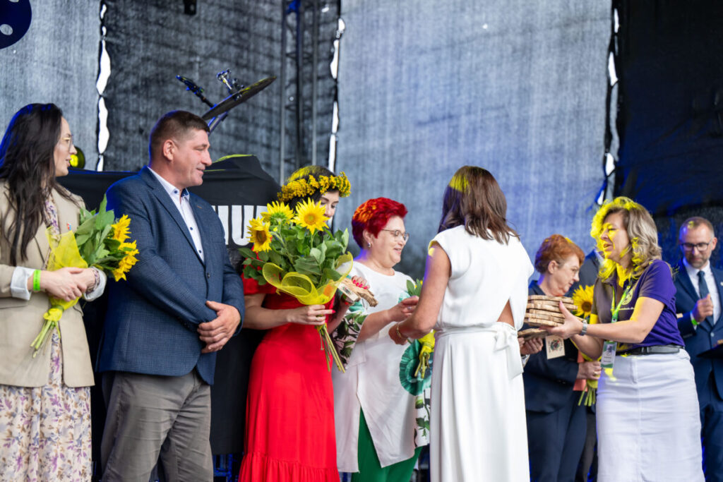 Grupa ludzi na scenie, wręczających sobie kwiaty i prezenty. W tle szare zasłony, dominują kolory zielony i żółty. Uroczysta atmosfera.