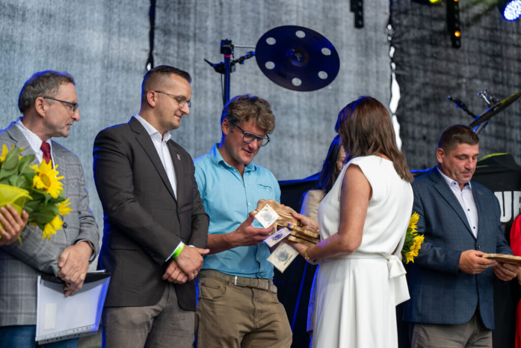Na zdjęciu grupa ludzi na scenie, wręcza nagrody. Mężczyzna w niebieskiej koszuli trzyma drewniane trofea. Tło ciemne, z elementami sprzętu muzycznego.