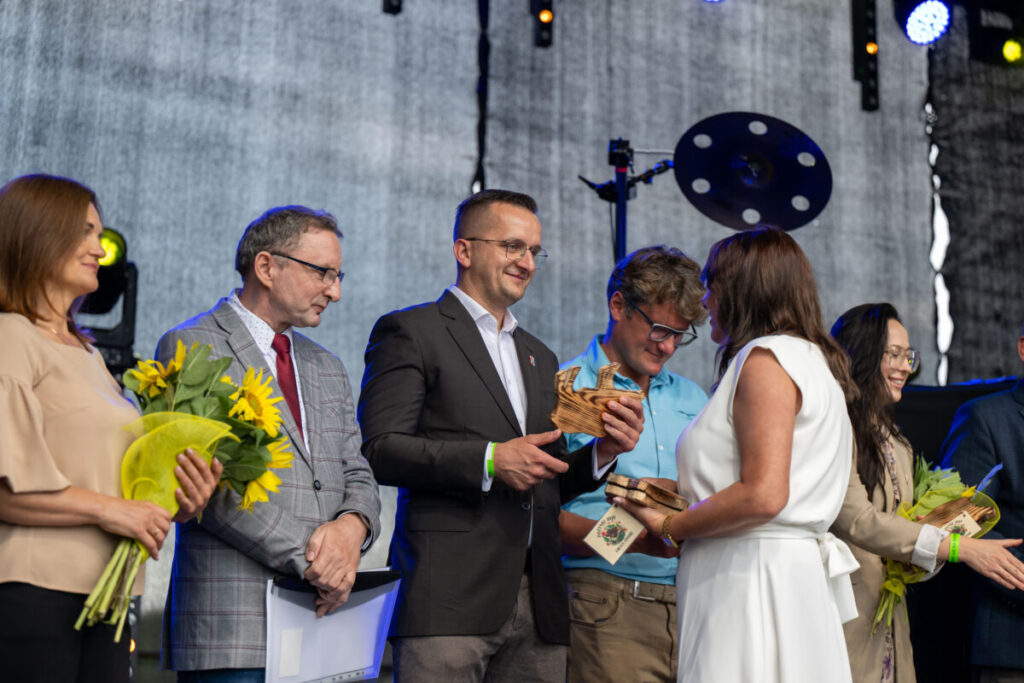 Na scenie odbywa się ceremonia wręczania nagród. Uczestnicy mają w rękach bukiety słoneczników i statuetki. Tło jest szare z kolorowymi światłami.