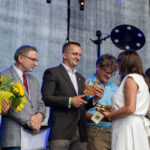 Na scenie odbywa się ceremonia wręczania nagród. Uczestnicy mają w rękach bukiety słoneczników i statuetki. Tło jest szare z kolorowymi światłami.