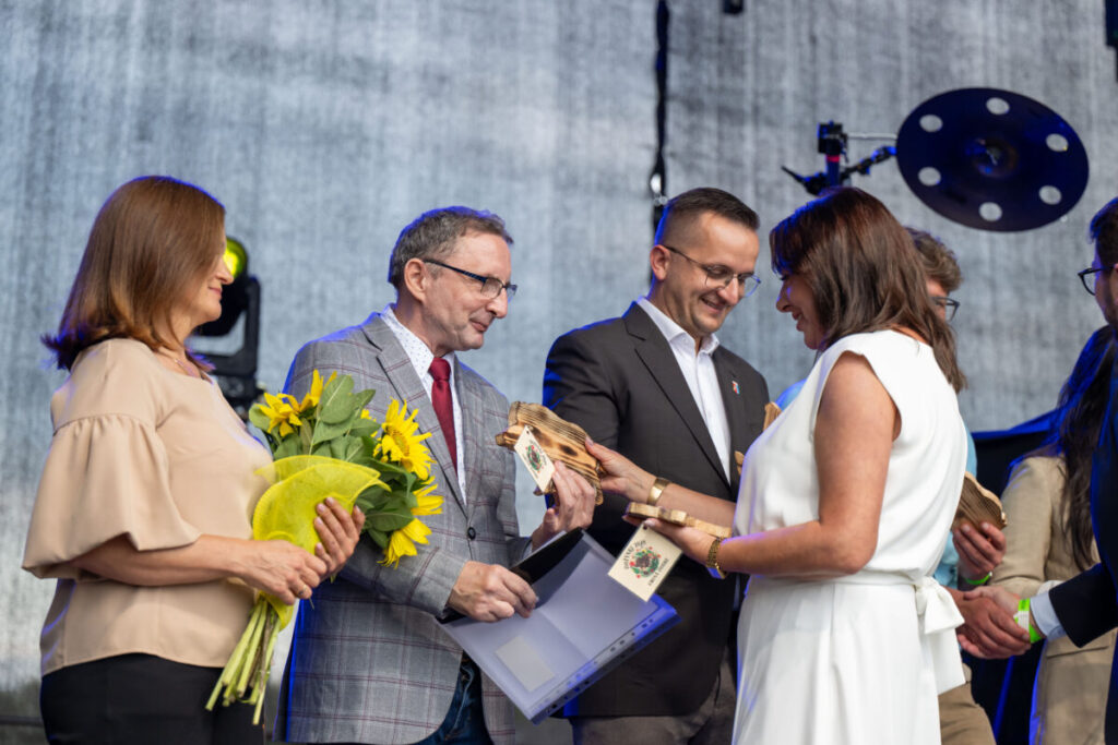 Grupa ludzi na scenie, trzyma kwiaty i nagrody. Mężczyzna w szarym garniturze i kobieta w bieli. Tło szare, scena.