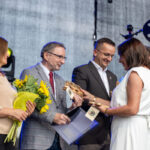 Grupa ludzi na scenie, trzyma kwiaty i nagrody. Mężczyzna w szarym garniturze i kobieta w bieli. Tło szare, scena.