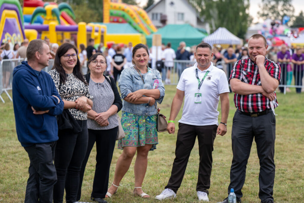Sześć osób stoi na trawie przed kolorowymi dmuchańcami. Tło to jasne namioty i ogrodzenie. Na zdjęciu dominują kolory: żółty i zielony.