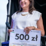 Kobieta w białej sukience trzyma wielki czek na 500 zł z motywem słoneczników. Stoi na scenie, w tle ciemne zasłony. Ma mikrofon w prawej dłoni.