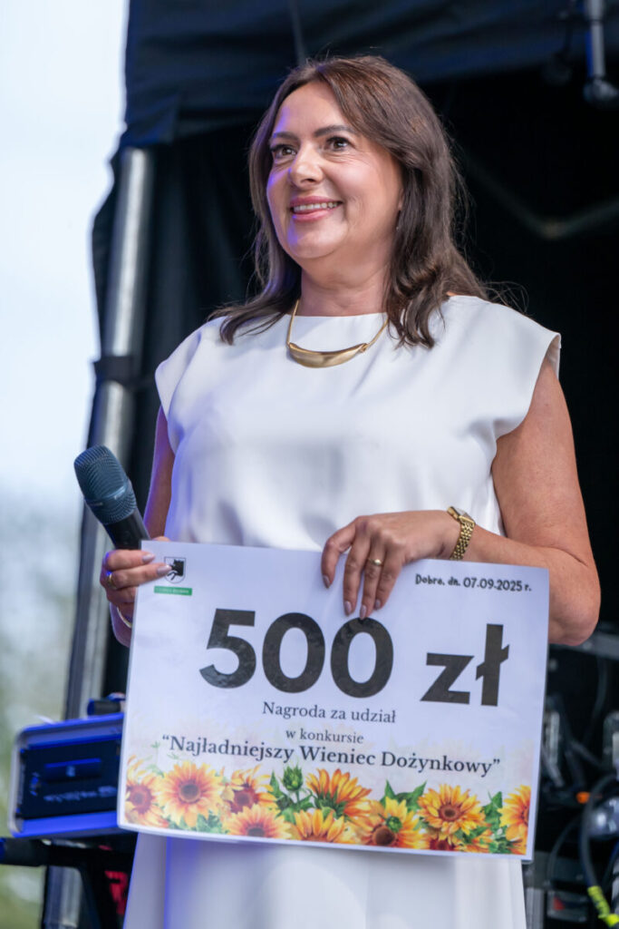 Kobieta w białej sukience trzyma wielki czek na 500 zł z motywem słoneczników. Stoi na scenie, w tle ciemne zasłony. Ma mikrofon w prawej dłoni.