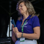 Kobieta w fioletowej koszulce, z dokumentami, stoi na tle ciemnego wnętrza ze sprzętem elektronicznym. Ma identyfikator na szyi.