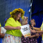 Na zdjęciu kobieta w zielonej koszulce odbiera nagrodę z napisem 500 zł. Ma wianek z żółtych kwiatów. Tło ciemne, z niebieskim oświetleniem.