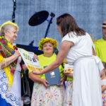 Na zdjęciu starsza kobieta w kolorowej chuście odbiera nagrodę od kobiety w bieli. W tle widoczni ludzie w żółtych koszulkach. Tło jest szare, sceniczne.