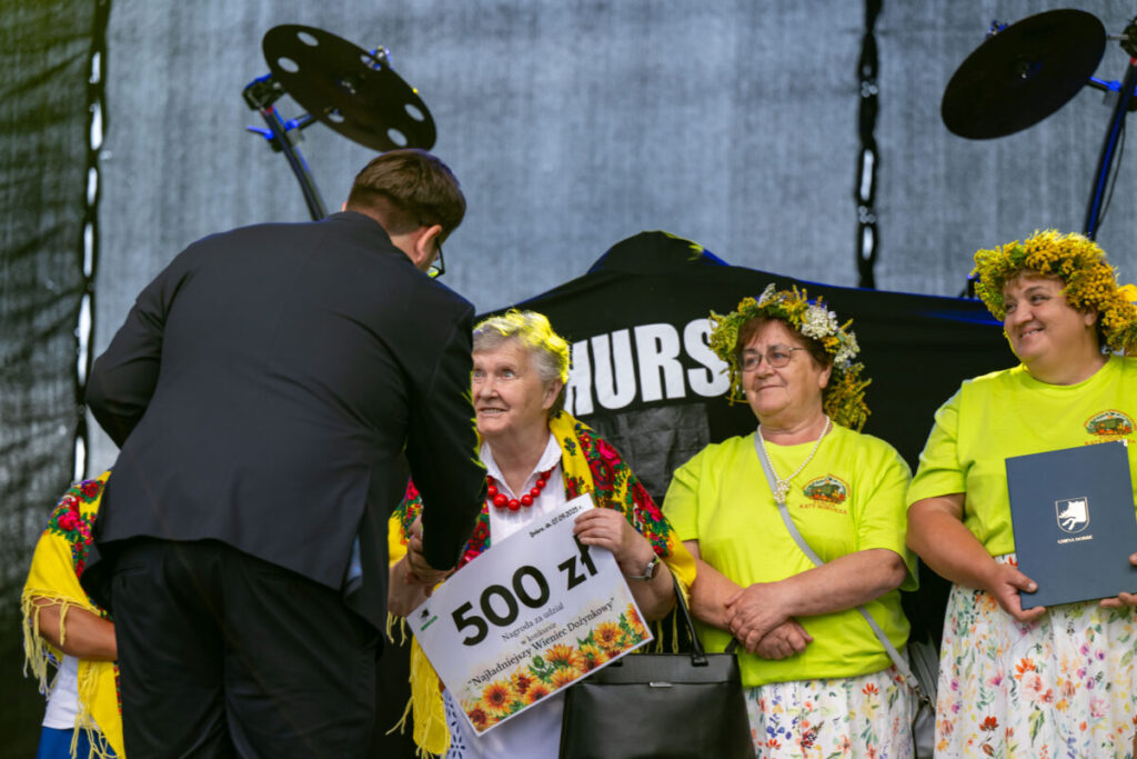 Grupa kobiet w wieńcach kwiatowych odbiera nagrodę na scenie. Starsza kobieta trzyma czek na 500 zł. Tło jest ciemne, sprzęt muzyczny z boku.