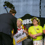 Grupa kobiet w wieńcach kwiatowych odbiera nagrodę na scenie. Starsza kobieta trzyma czek na 500 zł. Tło jest ciemne, sprzęt muzyczny z boku.