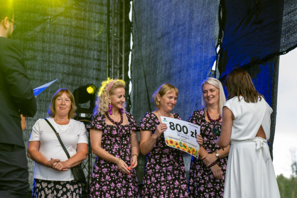 Na scenie stoją cztery kobiety, trzymają czek na 800 zł. Noszą sukienki w kwiaty, tło jest ciemne; światła sceniczne. Tło jest ciemne z efektem oświetlenia.