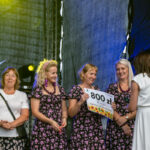 Na scenie stoją cztery kobiety, trzymają czek na 800 zł. Noszą sukienki w kwiaty, tło jest ciemne; światła sceniczne. Tło jest ciemne z efektem oświetlenia.